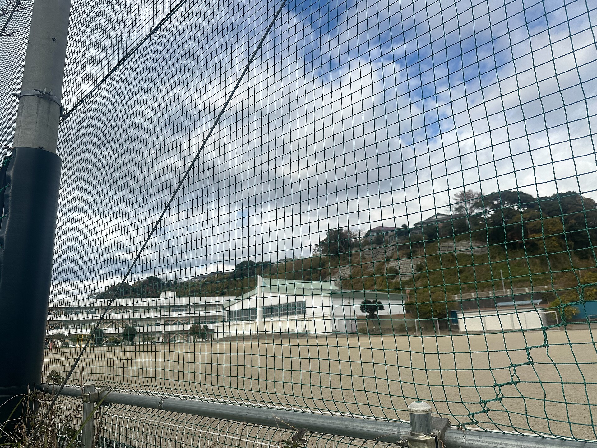 フェンス越しに見える校舎と体育館、緑の山を背にした校庭